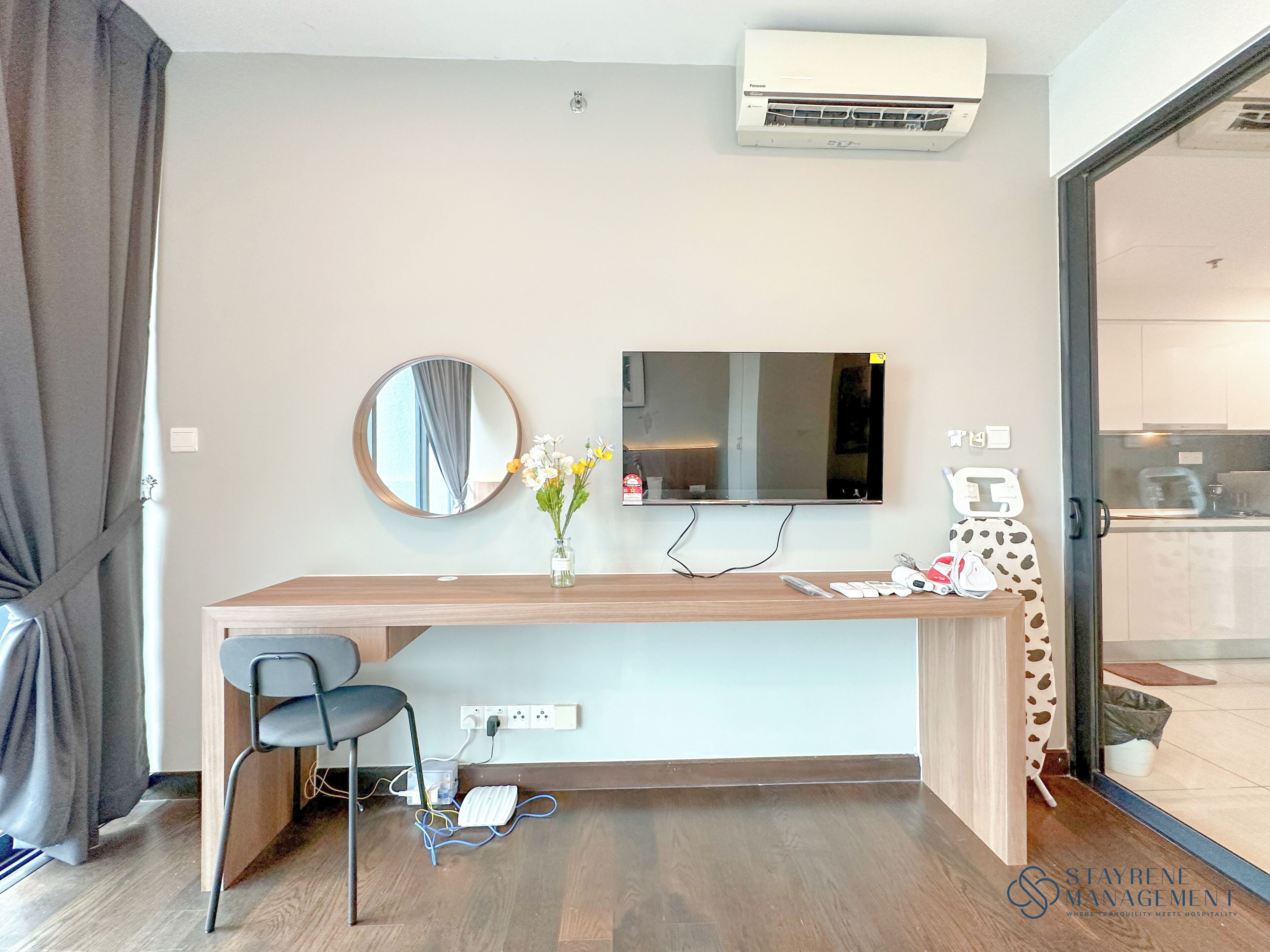 A modern hotel room with a wooden desk, a chair, a flat-screen TV, a mirror, and a vase with flowers.