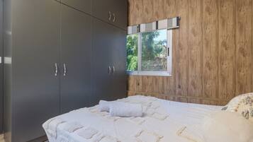 A bedroom with a large wardrobe, a bed with a white bedspread, and a window with a view of greenery.