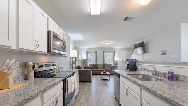 A modern kitchen with white cabinets, a microwave, and a coffee maker. There is a dining area with a sofa and a coffee table.