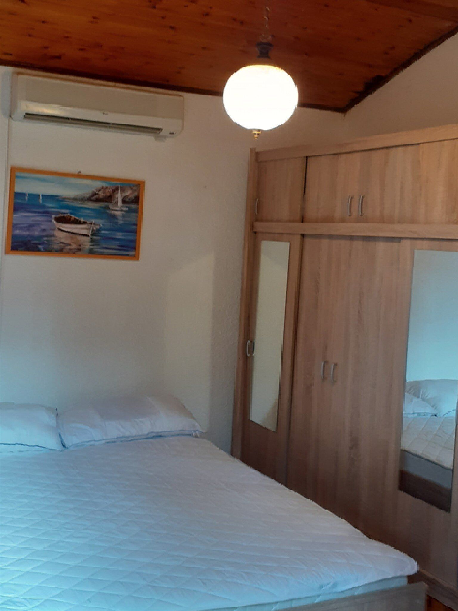 A bedroom with a bed, a wooden wardrobe, a painting of a boat on water, and a ceiling light.