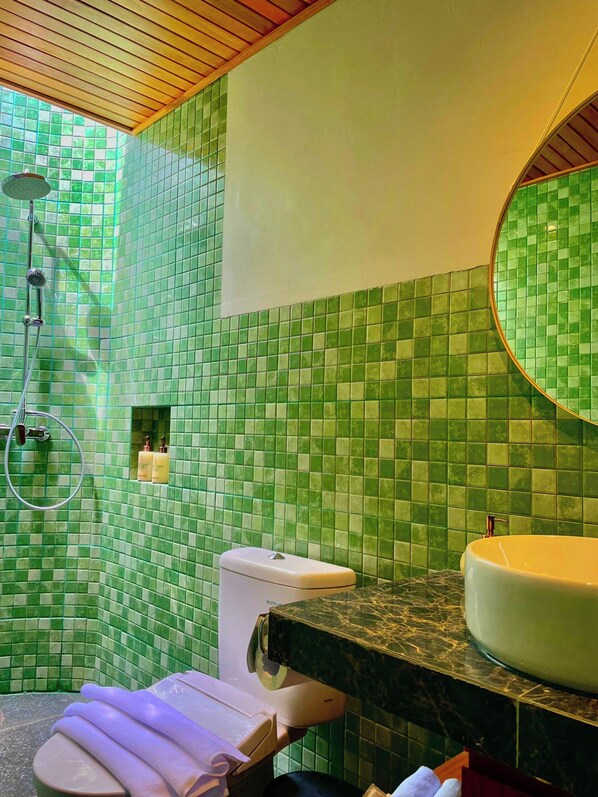 Un baño con mosaicos verdes, un inodoro blanco, un lavabo con encimera de mármol y una ducha con cabezal de ducha manual.
