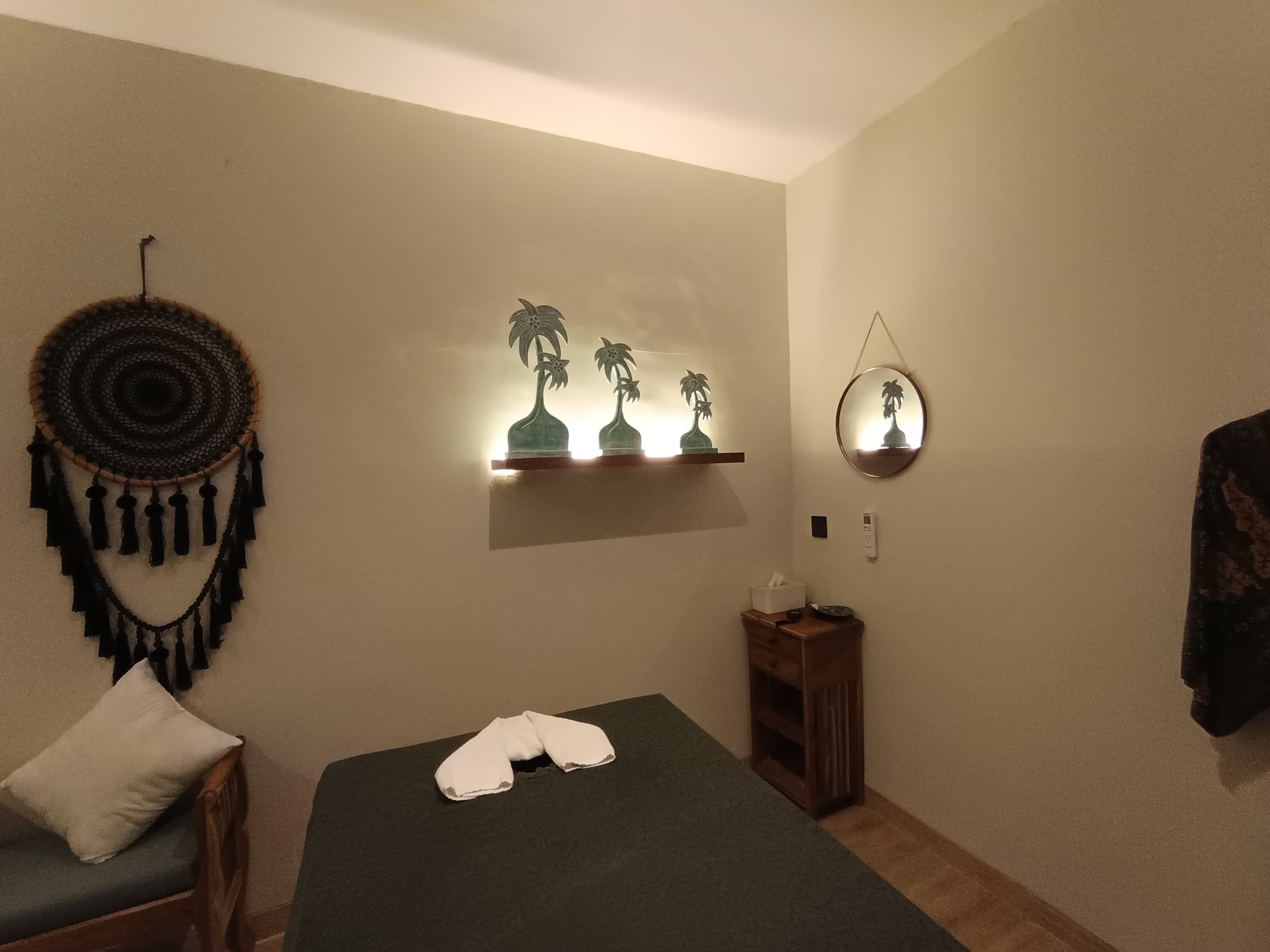 A room with a bed, a small wooden nightstand, a wall-mounted shelf with decorative items, a wall hanging, and a mirror.
