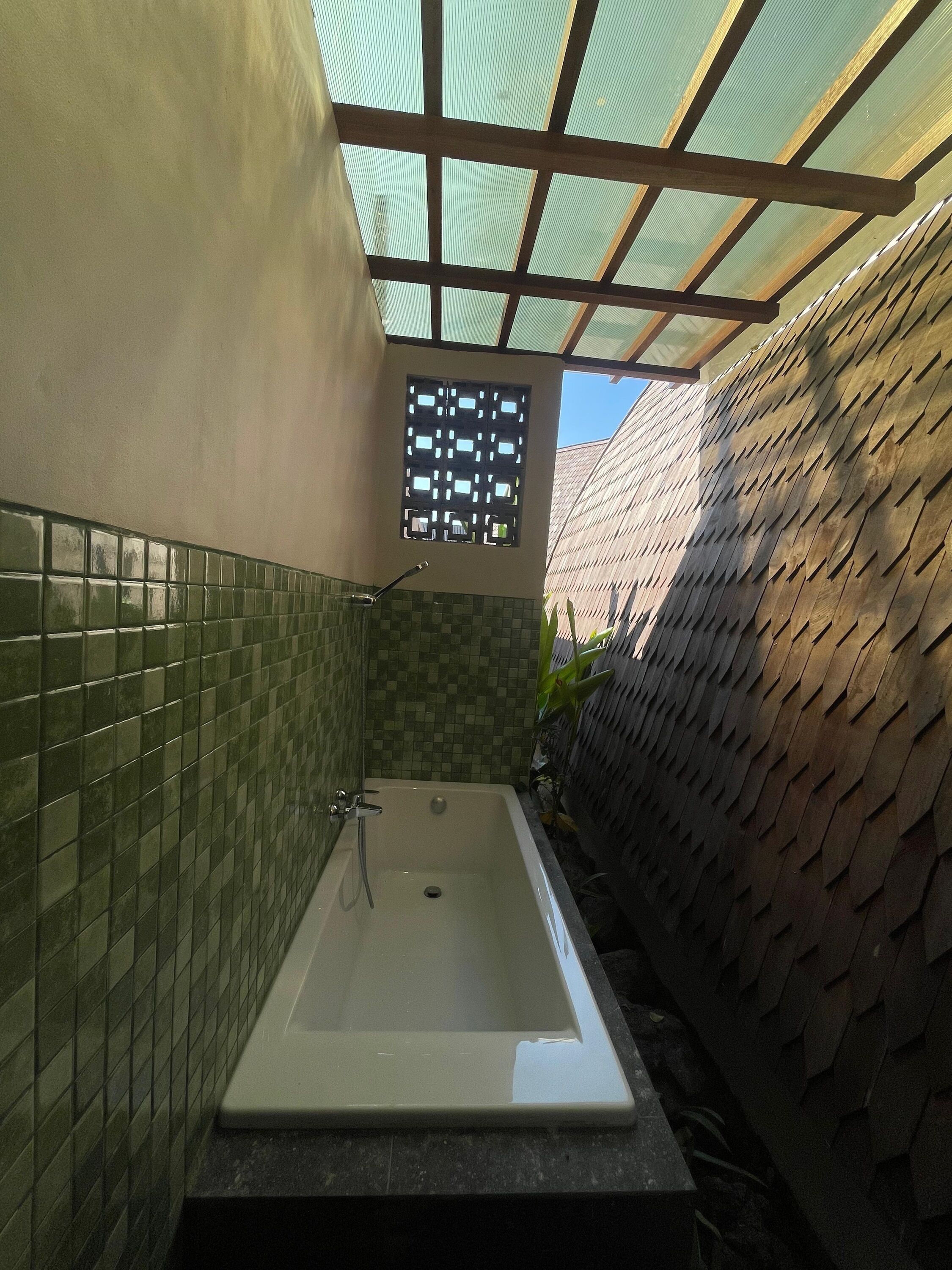 Ein Badezimmer mit freistehender Badewanne, gefliesten Wänden, einem Fenster mit geometrischem Muster und einer Decke mit lichtdurchlässiger Platte.