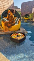 A woman sitting on a yellow and green woven chair by a pool, with a wicker basket of food and drinks in front of her.