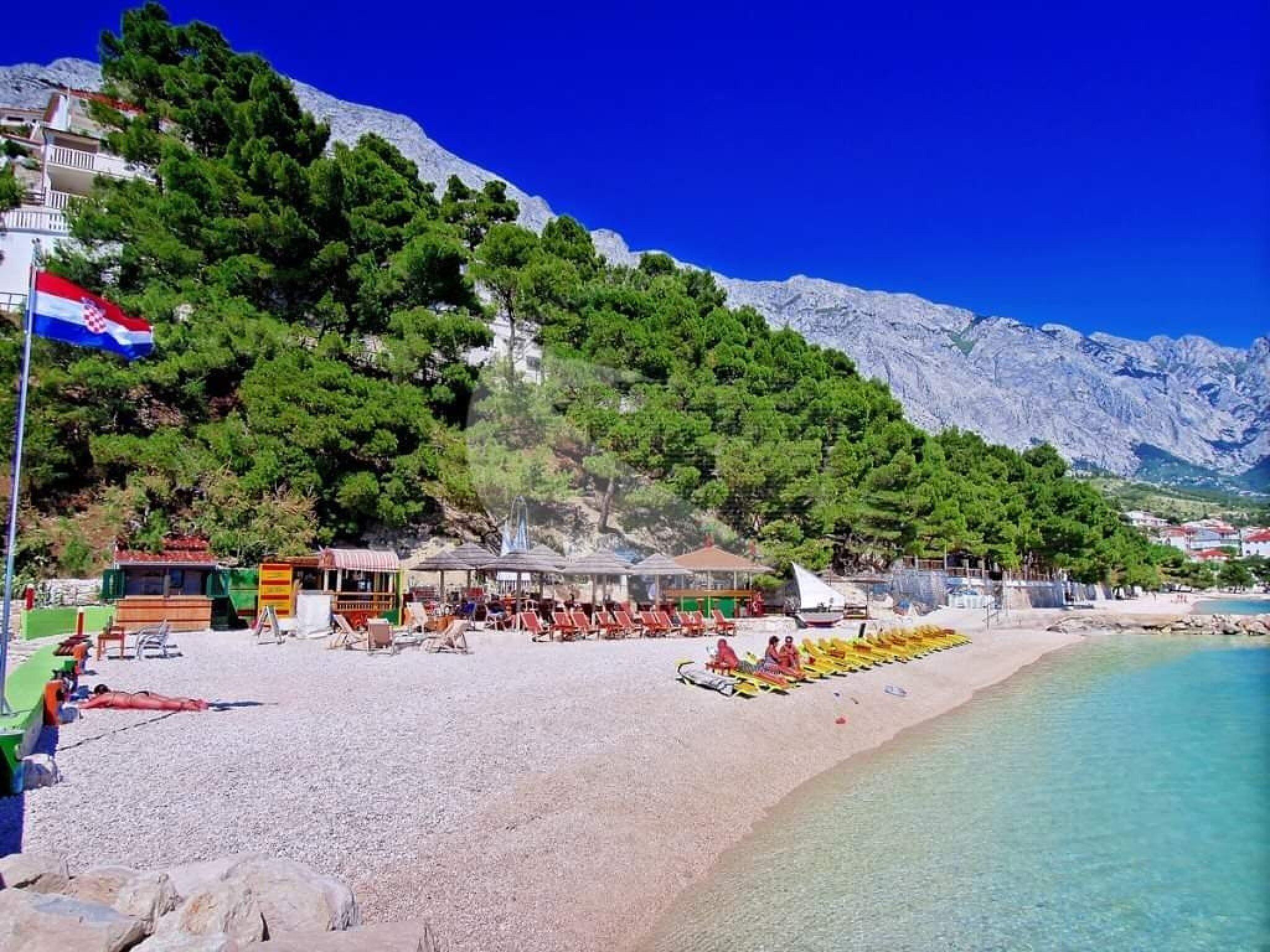Una platja amb aigua turquesa cristal·lina, una bandera i un paisatge de muntanyes al fons.
