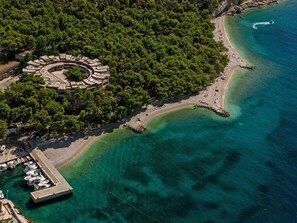 An aerial view of a coastal area with a circular stone structure surrounded by dense trees, a sandy beach, and clear blue waters. - Apartments Pezo (Baska Voda)