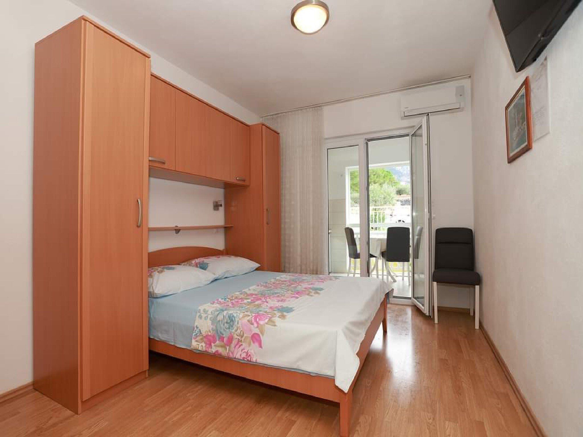 A bedroom with a bed, a wooden wardrobe, a small table with chairs, and a sliding glass door leading to an outdoor area.