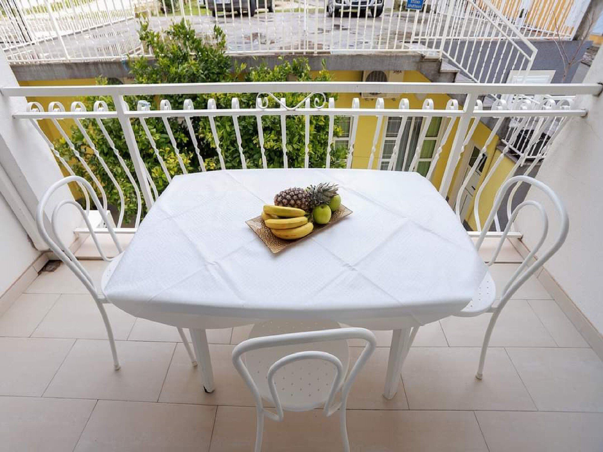 A balcony with a white table and chairs, a fruit bowl on the table, and a view of a street and greenery.