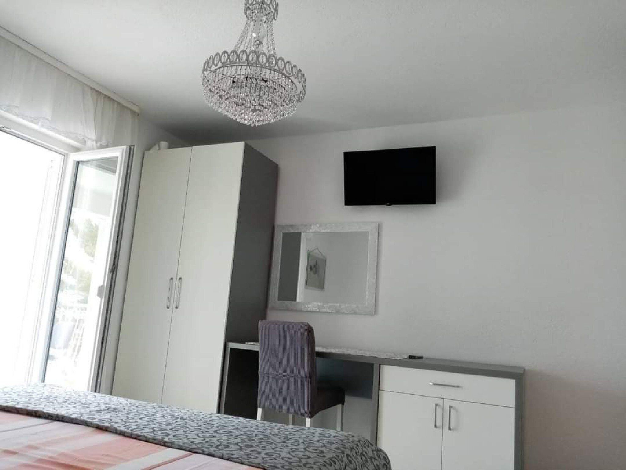 A bedroom with a bed, a wardrobe, a desk, a chair, a television, and a chandelier.