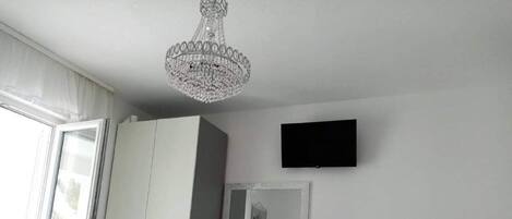 A bedroom with a bed, a wardrobe, a desk, a chair, a television, and a chandelier.