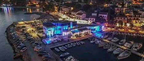 A coastal town at night with a brightly lit building with blue and purple lights, a marina with boats, and a densely packed residential area in the background.