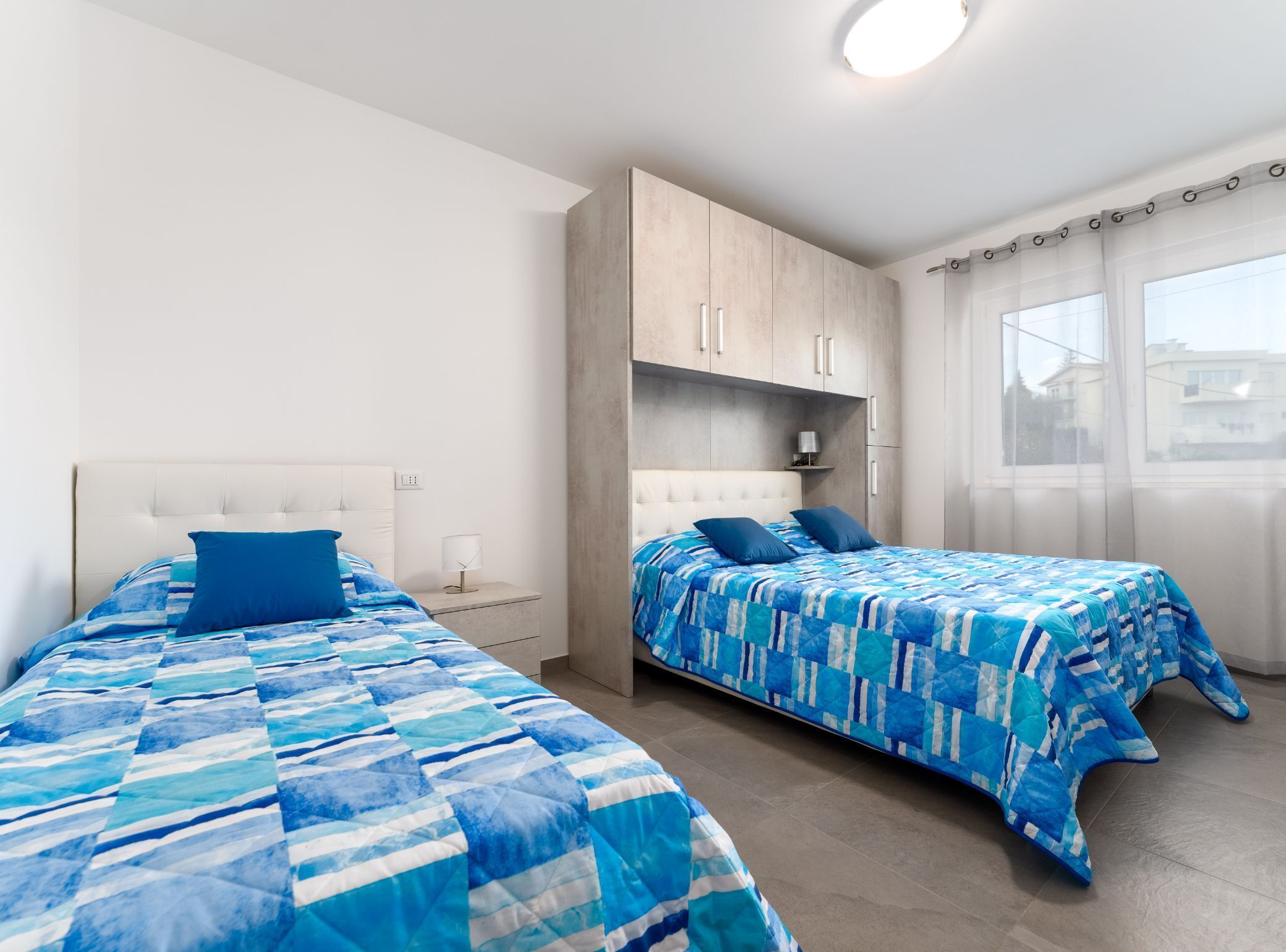 A bedroom with two beds, a wardrobe, a nightstand, and a window with curtains.