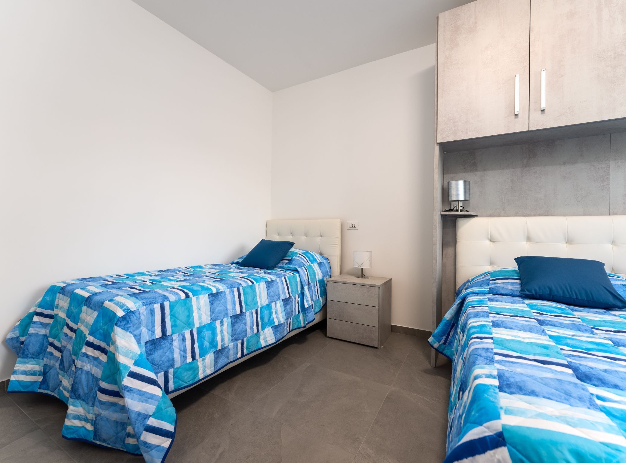 Een slaapkamer met twee bedden, elk voorzien van een blauw sprei, een nachtkastje met lamp, en een garderobe met grijze afwerking.