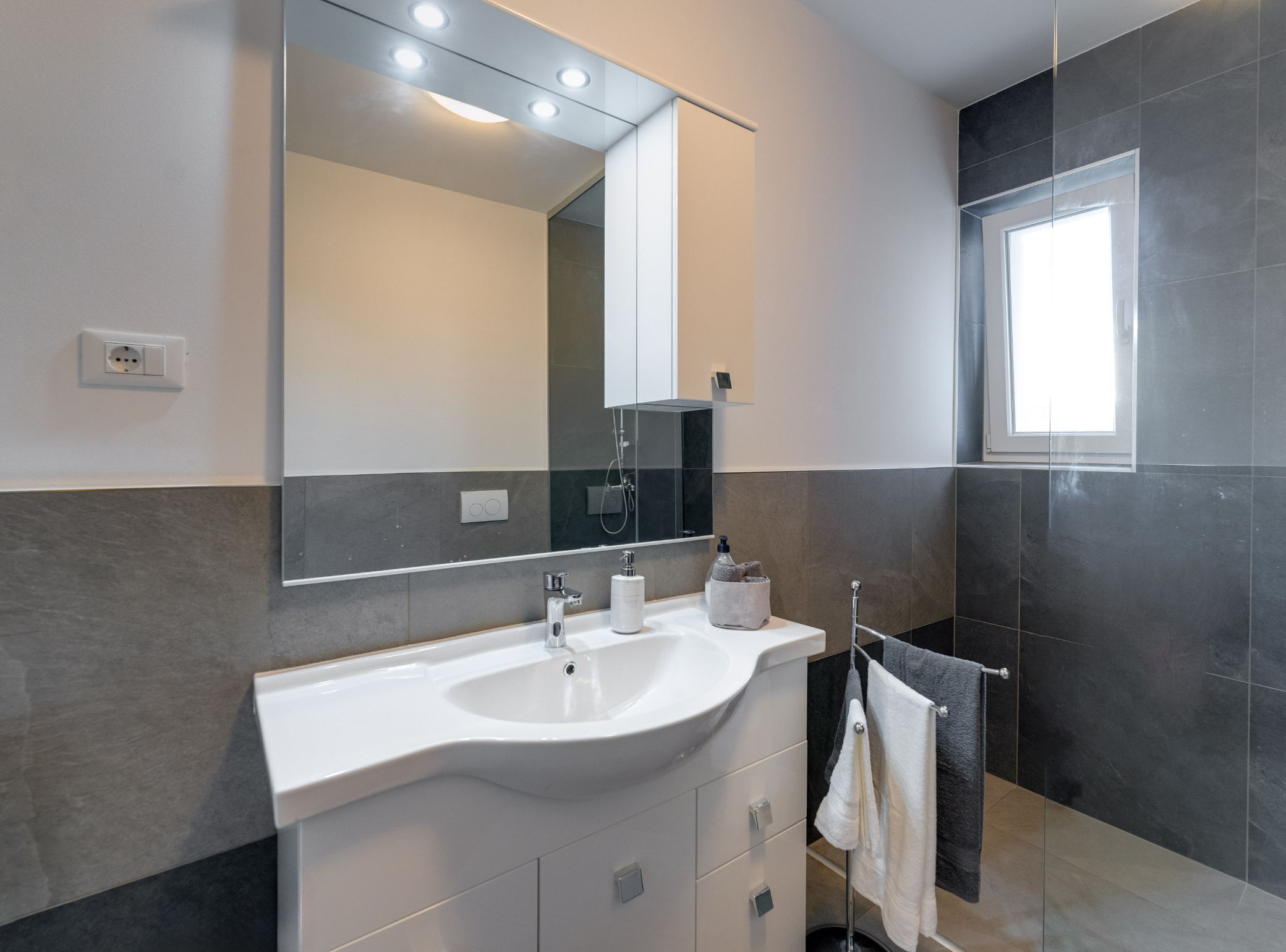 A modern bathroom with a large mirror, white sink, and dark tiled walls.