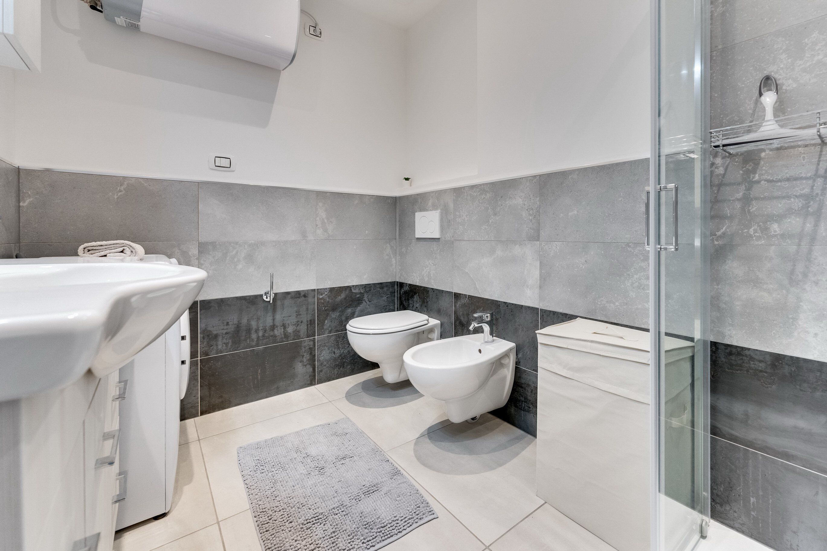 A modern bathroom with a glass shower enclosure, a white sink, two toilets, and a marble-tiled wall.