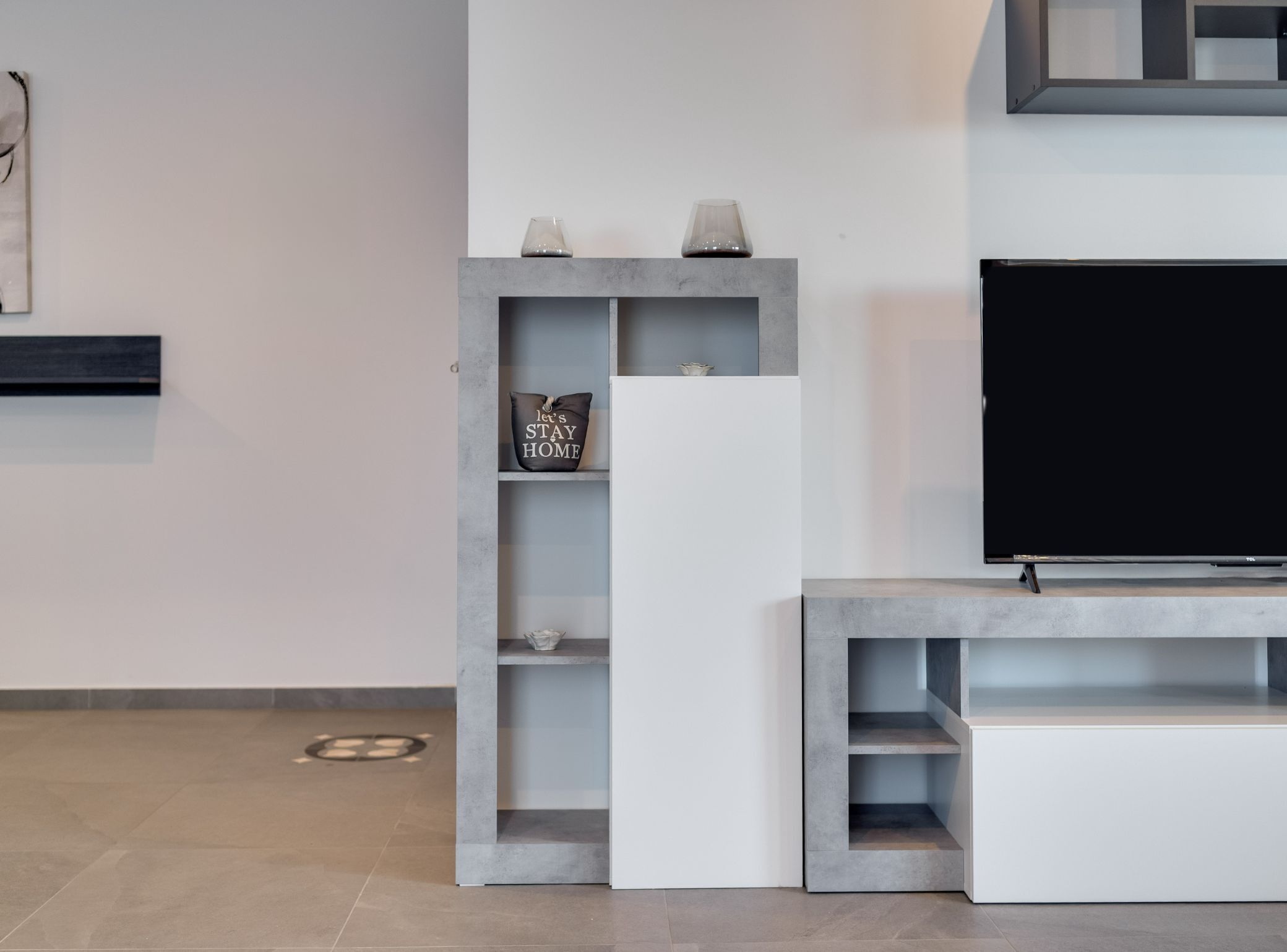 A modern living room with a flat-screen TV on a stand, grey shelving units, and a minimalist decor style.