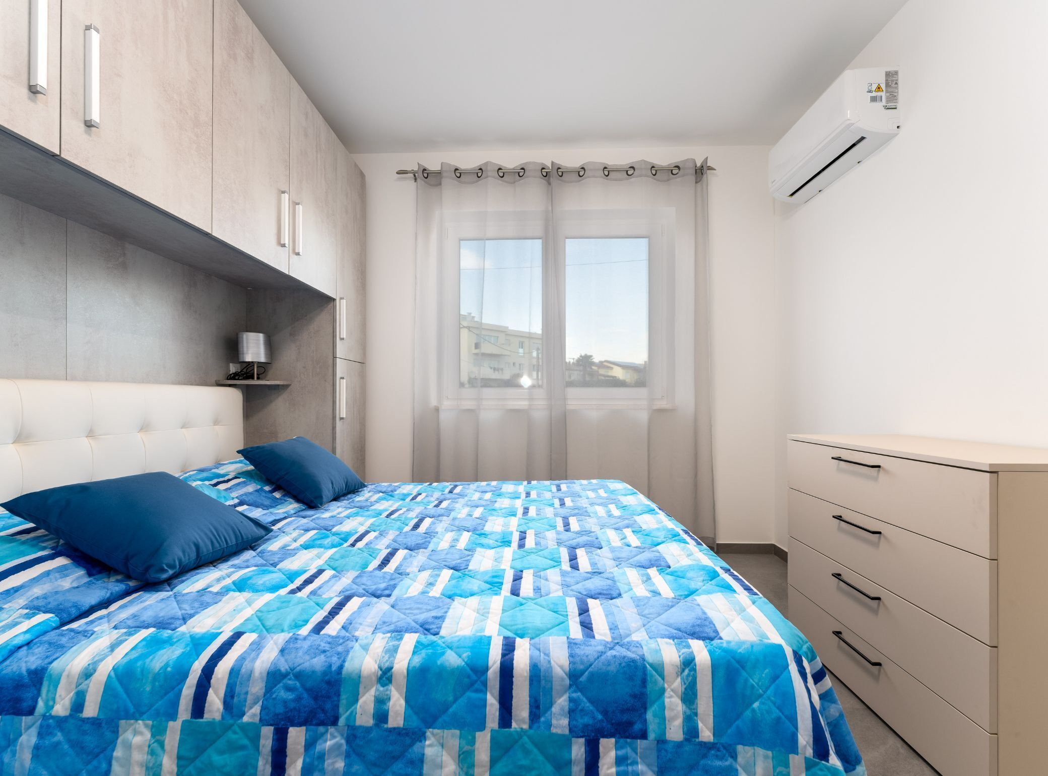 A bedroom with a bed, a wardrobe, a window with curtains, and an air conditioning unit.
