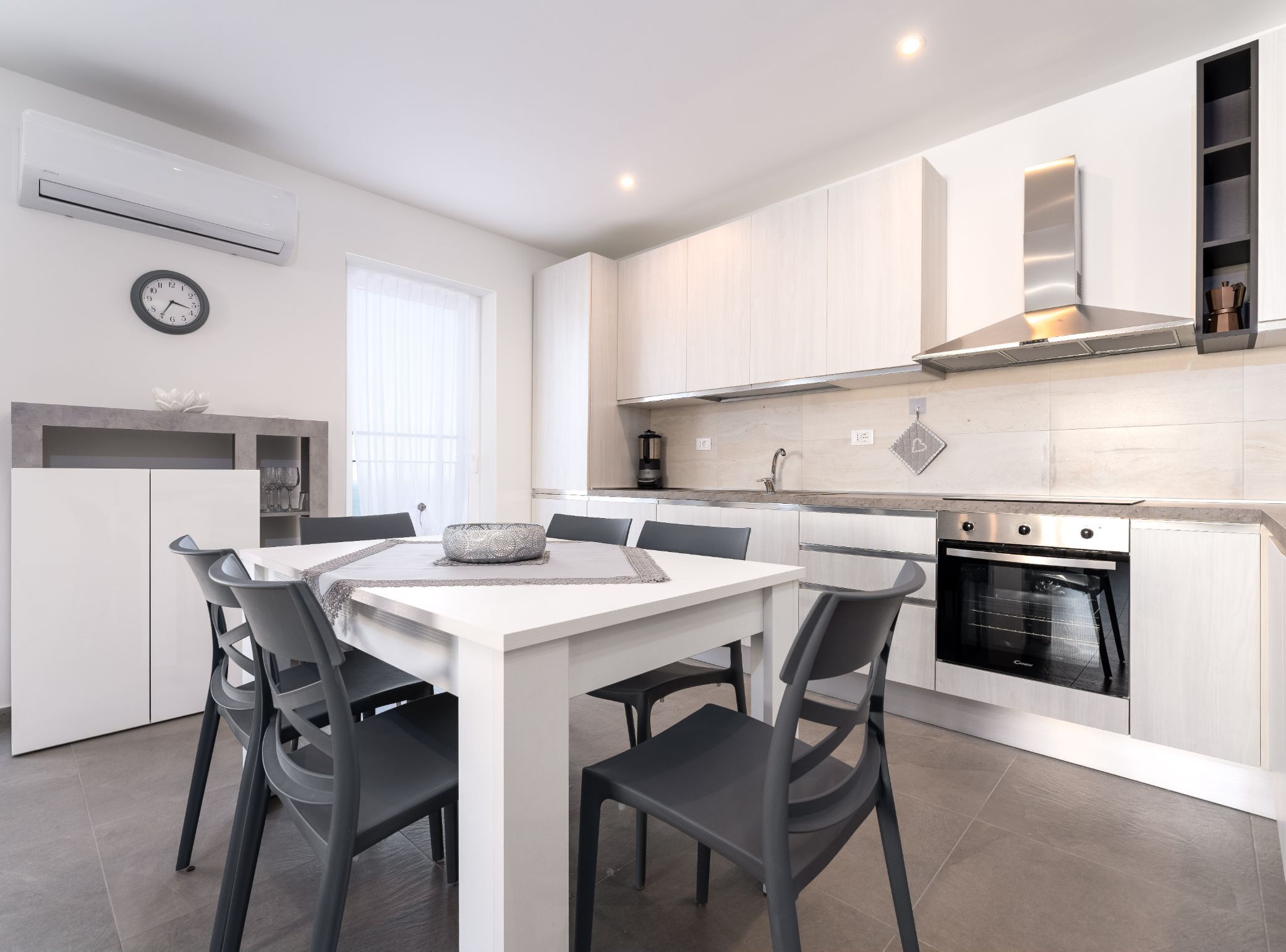Una cocina moderna con armarios blancos, horno empotrado y comedor con mesa y sillas blancas.
