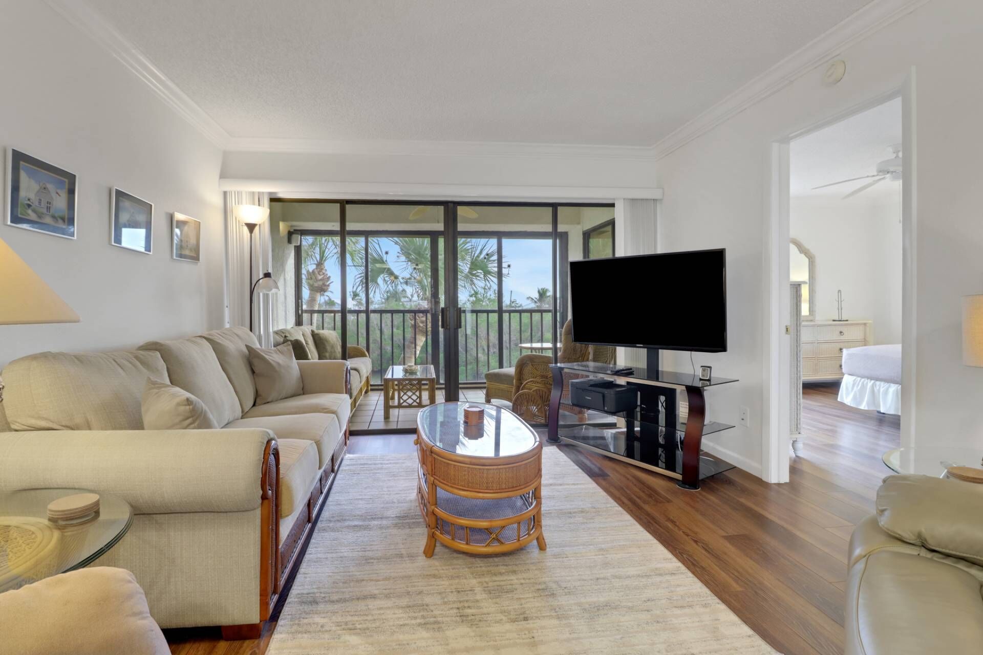 A living room with a sofa, a glass coffee table, a television on a stand, and a balcony with a view of palm trees.