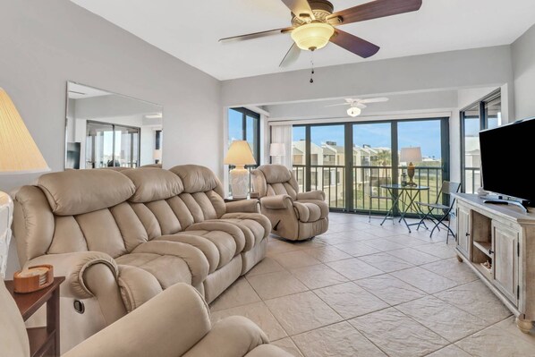 A spacious living room with a large beige sofa, a ceiling fan, and a balcony with a table and chairs.