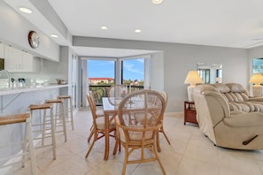 A modern kitchen with white cabinets, a dining area with wicker chairs, a sofa, and a view through the glass doors. - Lovely End-Unit, Lagoon-Front Community, South of Mid-Island! Heated Community Pool! Free Parking! (Fort Myers Beach)