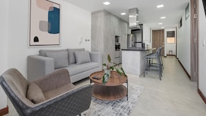Una sala moderna con un sofá gris, un sillón negro y una mesa de centro de madera. Se observa una cocina con estufa y refrigerador al fondo.