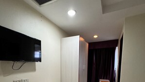 Een hotelkamer met een flatscreen televisie aan de muur, een klein nachtkastje, een uitschuifbed, een raam met gordijnen en een betegelde vloer.