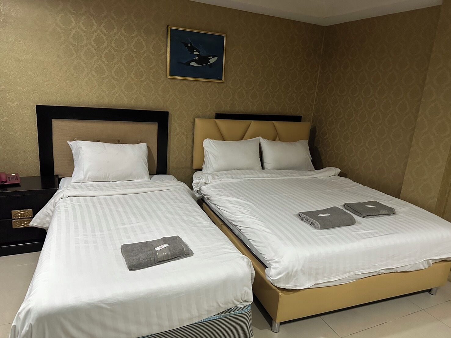 Habitación de hotel con dos camas, cabeceros color beige, ropa de cama blanca, un cuadro en la pared y una mesita de noche con un teléfono y un objeto decorativo.