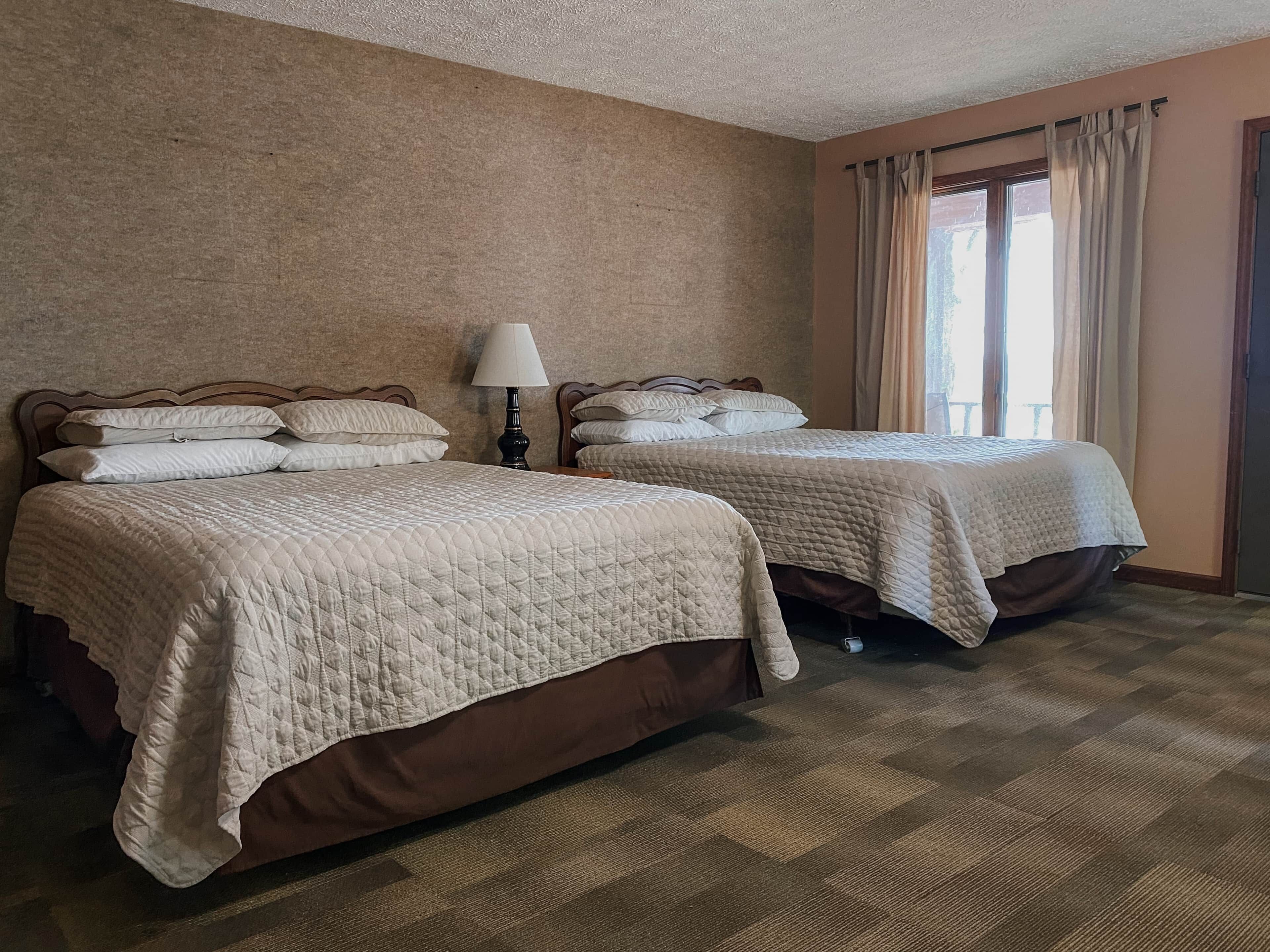 Two queen beds with matching headboards, a bedside lamp, and a window with curtains.