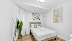 Un dormitorio moderno con cama de madera, ropa de cama blanca, una mesita de noche y una planta en maceta, junto con un cuadro abstracto en la pared.