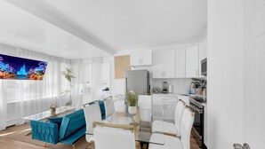 Una sala moderna con una mesa de comedor de vidrio, sofás blancos y una cocina con gabinetes y electrodomésticos blancos.