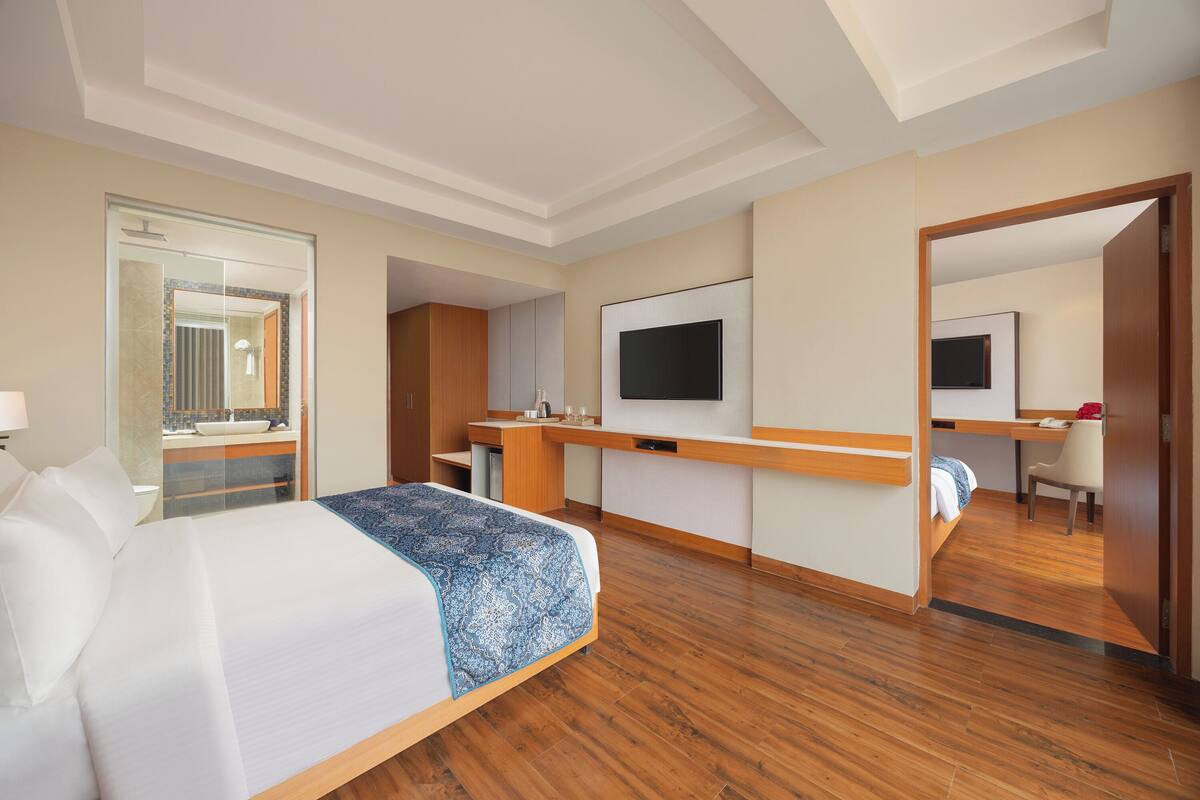 A hotel room with a bed, a desk, a TV, and a bathroom visible through a glass door.