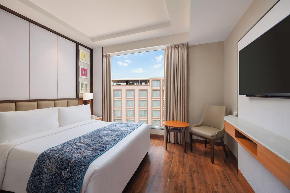 A hotel room with a large bed, a desk, a chair, a TV, and a window with a view of buildings.