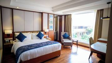 Quarto de hotel com uma cama grande, duas cadeiras, uma mesinha e vista para a cidade.