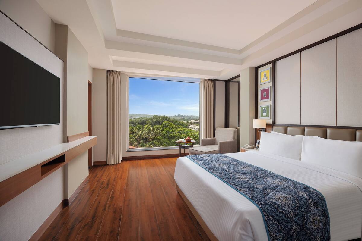A hotel room with a large bed, a TV, a small table, and a view of greenery outside.