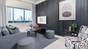 A modern living room with a grey sofa, a colorful patterned sofa, a round coffee table, and a black TV stand. There is a cityscape view through the window, and a large abstract painting on the wall.