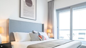 A modern hotel room with a large bed, a headboard, two bedside tables with lamps, a window with curtains, and a balcony with chairs.
