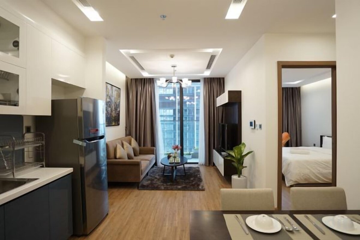 Et moderne hotelværelse med tekøkken, opholdsområde med sofa og sofabord, spiseplads med bord dækket til to personer, og et stort vindue med udsigt over byen.