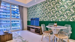 Ein modernes Wohnzimmer mit großem Fenster, Flachbildfernseher, Esstisch mit Stühlen und einer grünen Wand mit Blätermotiv.
