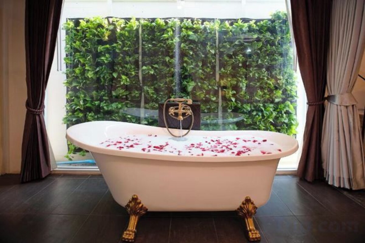 A freestanding bathtub with rose petals, positioned in front of a window with a view of greenery.
