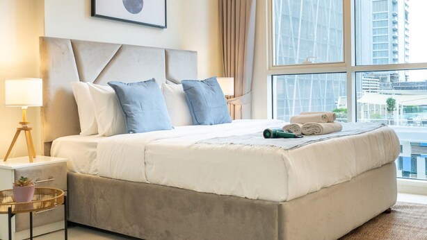 A modern hotel room with a large bed, a headboard, a bedside table with a lamp, and a view of the city through large windows. - 2bed Chic Apt Burj Khalifa Gym/pool by Belvilla (Dubai)