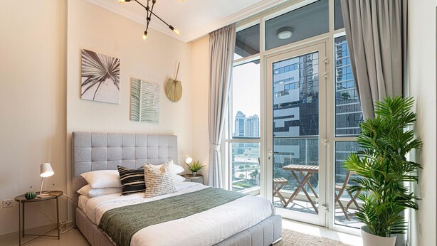 A modern bedroom with a large bed, a view of the city through floor-to-ceiling windows, and a balcony with a potted plant. - Apartment in Downtown Dubai Near Burj Khalifa (Dubai)