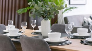 A dining area with a wooden table set for a meal, surrounded by grey chairs. A white vase with green foliage is placed on the table.
