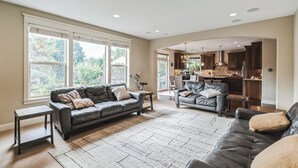 Un vaste salon avec un canapé en cuir foncé, une table basse et un tapis. On y trouve également un coin cuisine avec des meubles en bois et une table à manger, visibles à travers une porte ouverte.