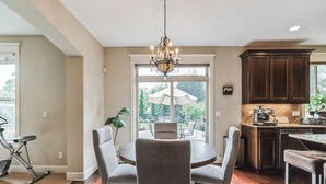 Un espace repas agrémenté d’un lustre, d’une table ronde et de chaises. On y trouve également une cuisine avec des meubles de couleur sombre et un plan de travail.