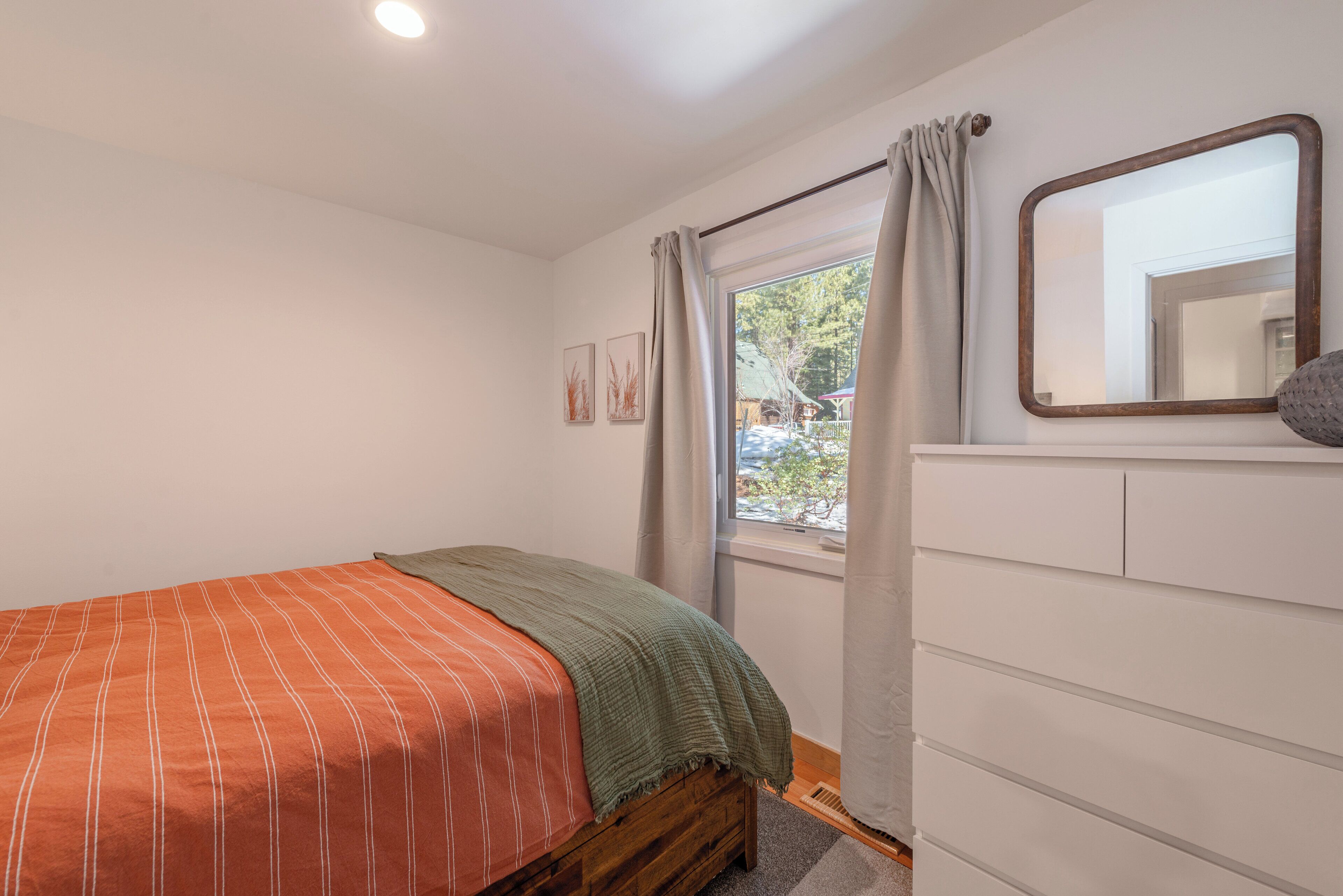 A bedroom with a bed, a dresser, a window with curtains, and a mirror on the wall.