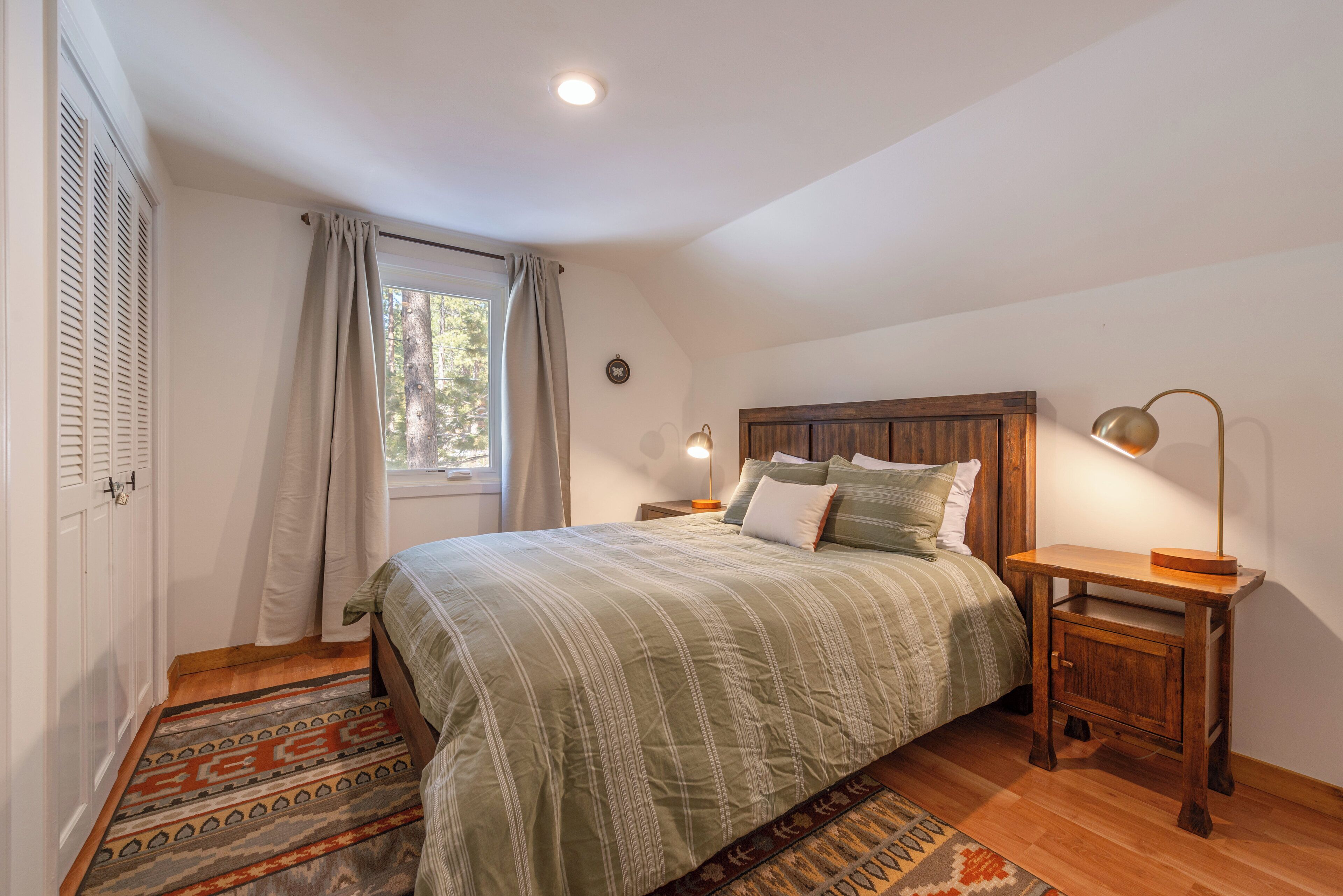 A bedroom with a bed, a nightstand, a lamp, a window with curtains, and a door with shutters.