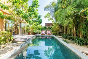 A swimming pool with two red and white striped lounge chairs, surrounded by lush greenery and a wooden building. - Hygge Villa Ubud (Ubud)