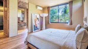 A modern bedroom with a large bed, a window with curtains, a bedside lamp, and a bathroom visible through an open door. - Hygge Villa Ubud (Ubud)