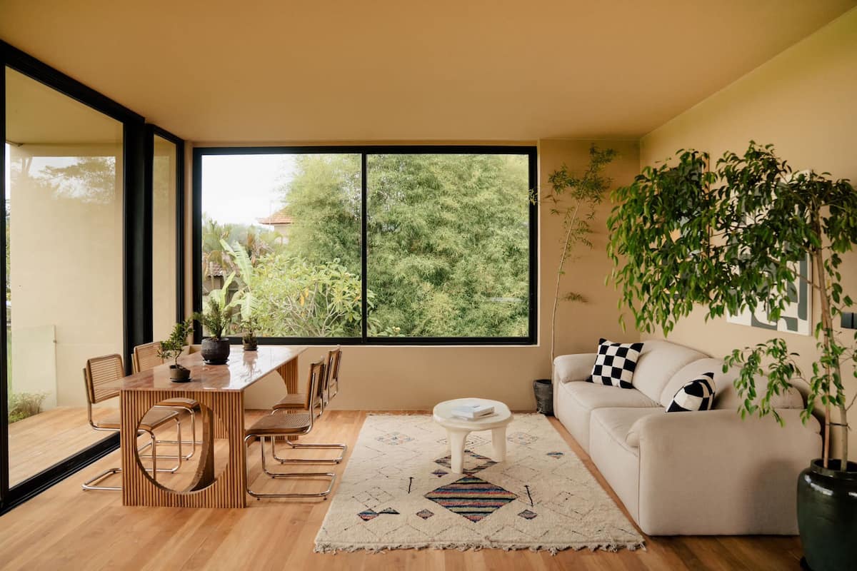 Una sala de estar moderna con una mesa de comedor de madera, sofá blanco y amplios ventanales con vistas a zonas verdes.